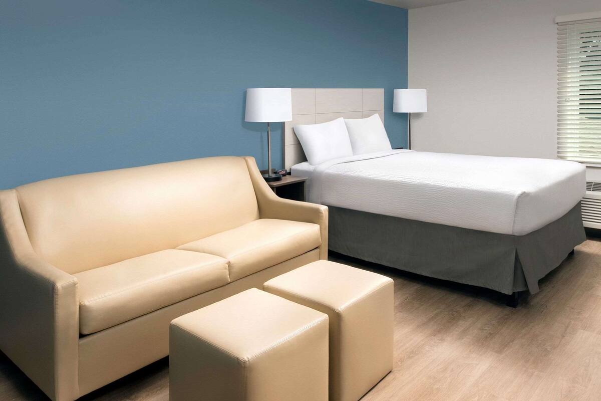 A hotel room with a beige sofa, a matching ottoman, a bed with white linens, and two bedside lamps.