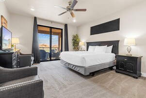 A hotel room with a large bed, a ceiling fan, a desk with a lamp, a chair, and a view of the outdoors.
