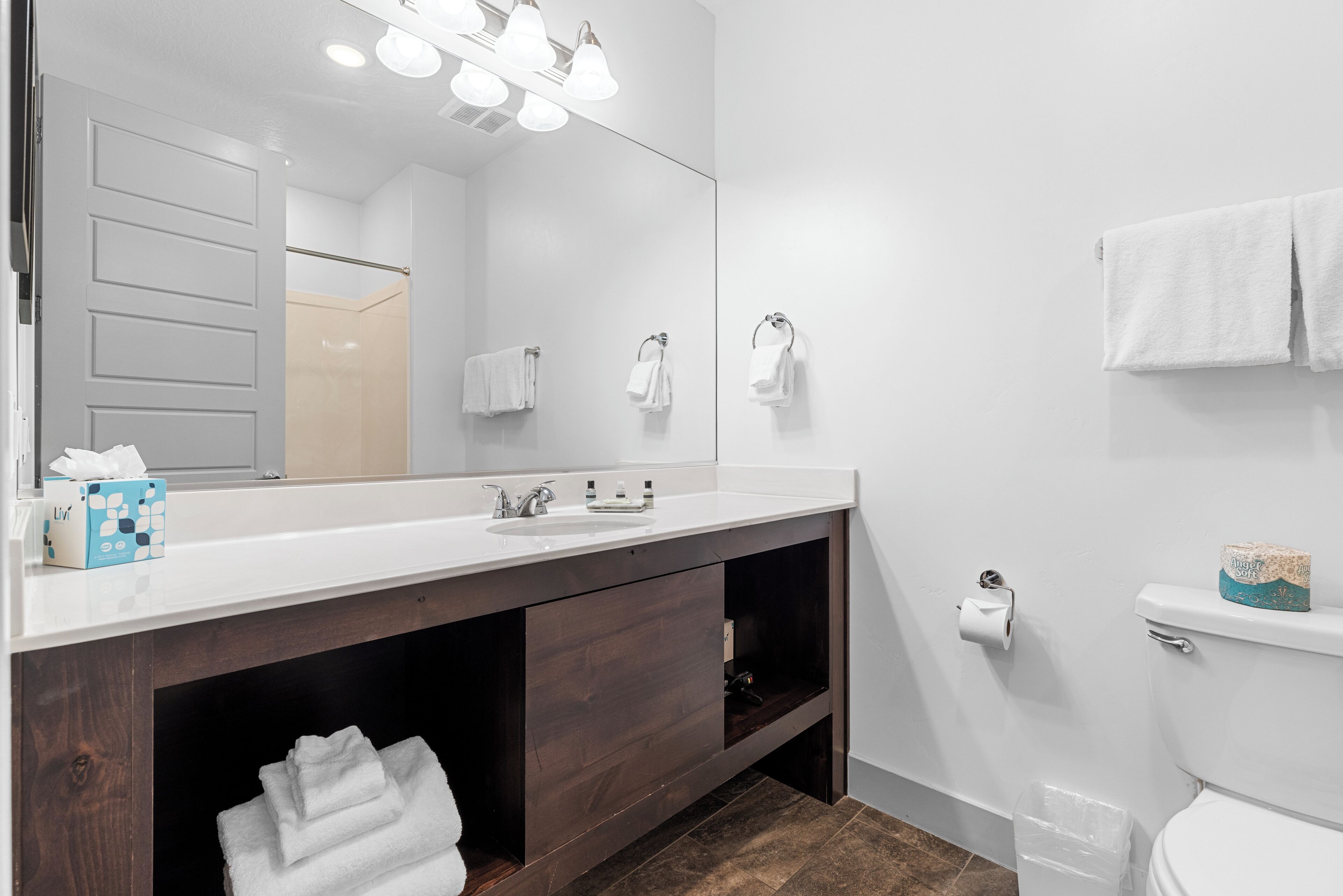 A modern bathroom with a white vanity, dark wooden cabinets, a large mirror, and a toilet with a paper towel dispenser.