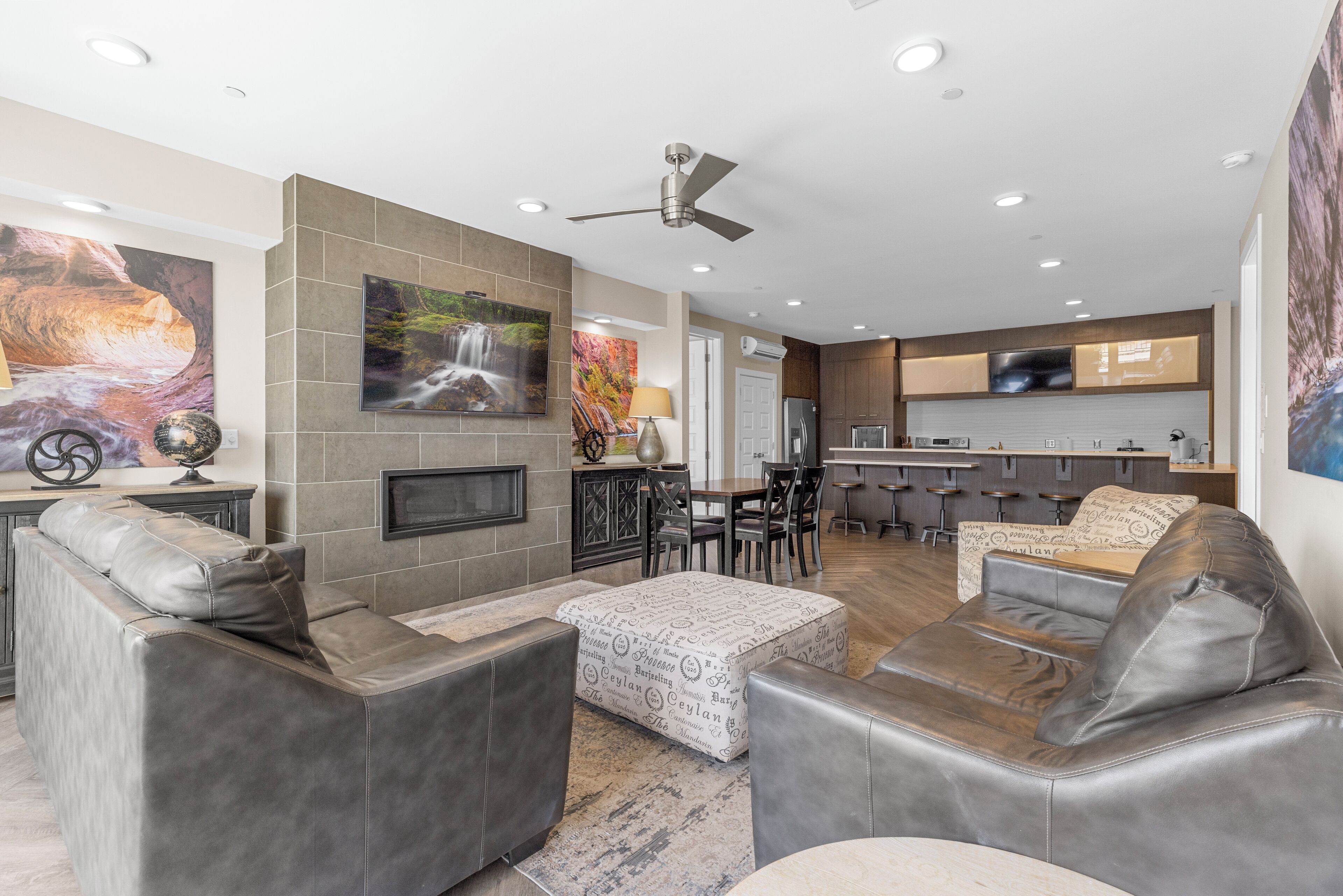 A modern living room with leather sofas, a coffee table, and a fireplace. There is a dining area with a bar and kitchen in the background.