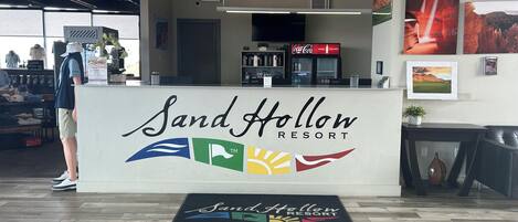 Un comptoir d’accueil se trouve au Sand Hollow Resort, avec une enseigne au-dessus, un réfrigérateur Coca-Cola et une vue sur le paysage par la fenêtre.