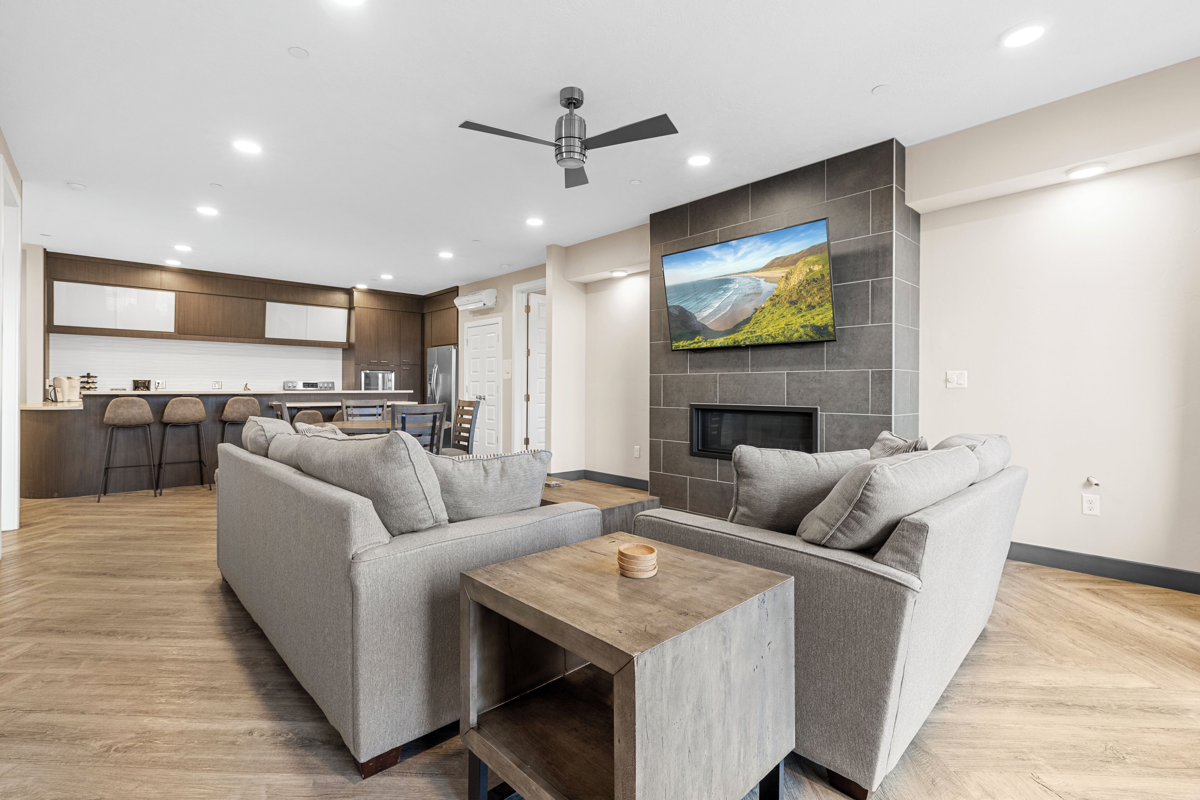A modern living room with a grey sectional sofa, a wooden coffee table, and a flat-screen TV mounted on the wall above a fireplace.