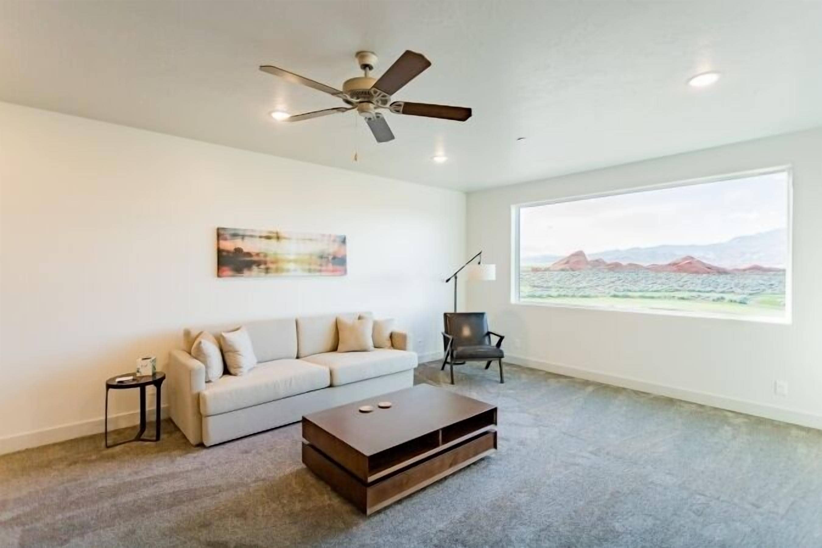 A modern living room with a white sofa, a coffee table, a ceiling fan, a floor lamp, and a large window with a mountain view.