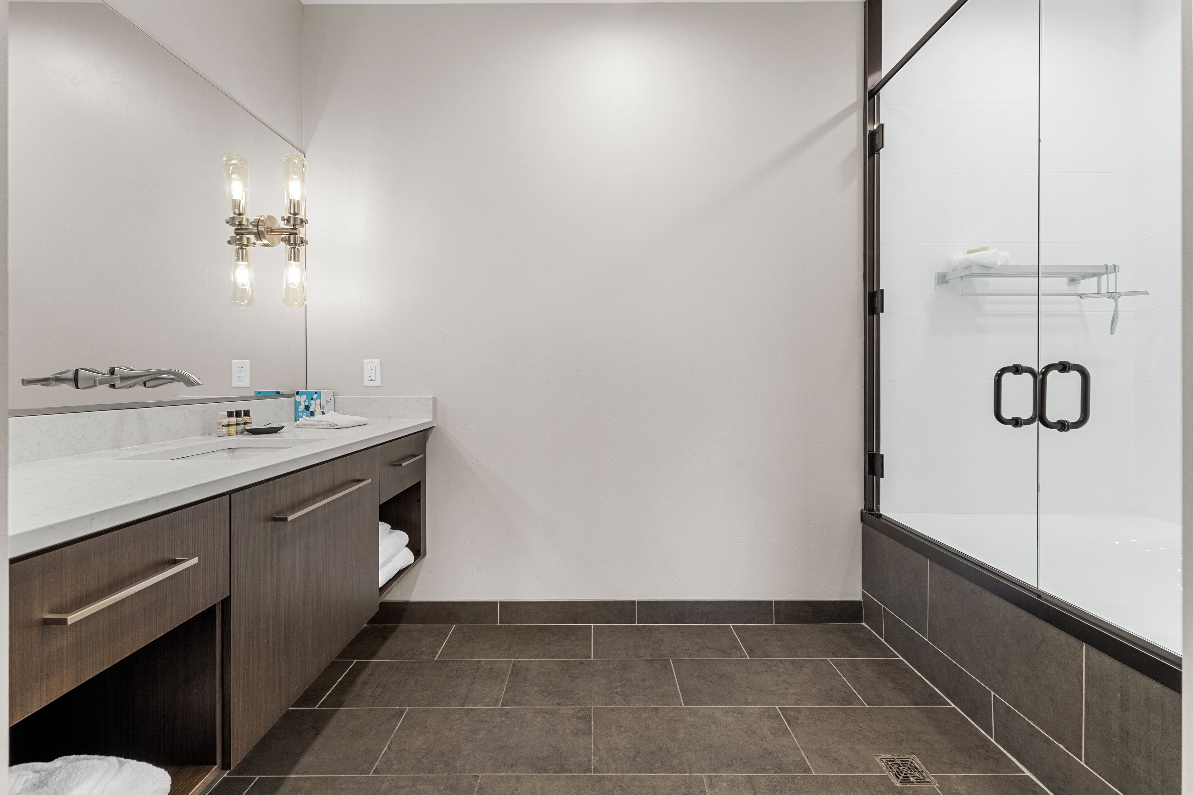 Un baño moderno con un espejo grande, una ducha con puertas de vidrio y piso cubierto de azulejos blancos.