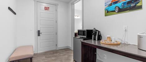 Un moderno vestíbulo de hotel con una puerta de salida, un banco, una kitchenette equipada con microondas, cafetera y una bandeja con tazas.