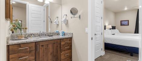 A bathroom with a wooden vanity, a large mirror, and a sink. There is a bed with a blue mattress in the adjacent room.