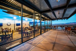 An outdoor dining area with glass walls, wooden tables, and chairs, overlooking a mountainous landscape.
