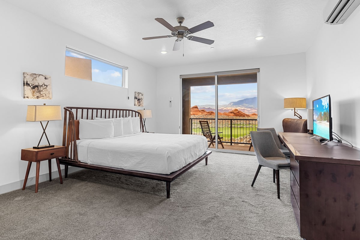 A bedroom with a bed, desk, chair, and a balcony with a view of the desert.