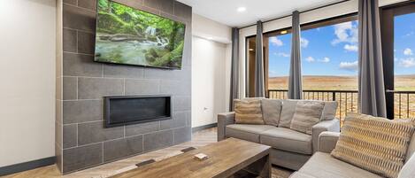 A modern living room with a flat-screen TV mounted on a stone wall, a grey sofa with patterned cushions, a wooden coffee table, and large windows offering a view of a landscape.