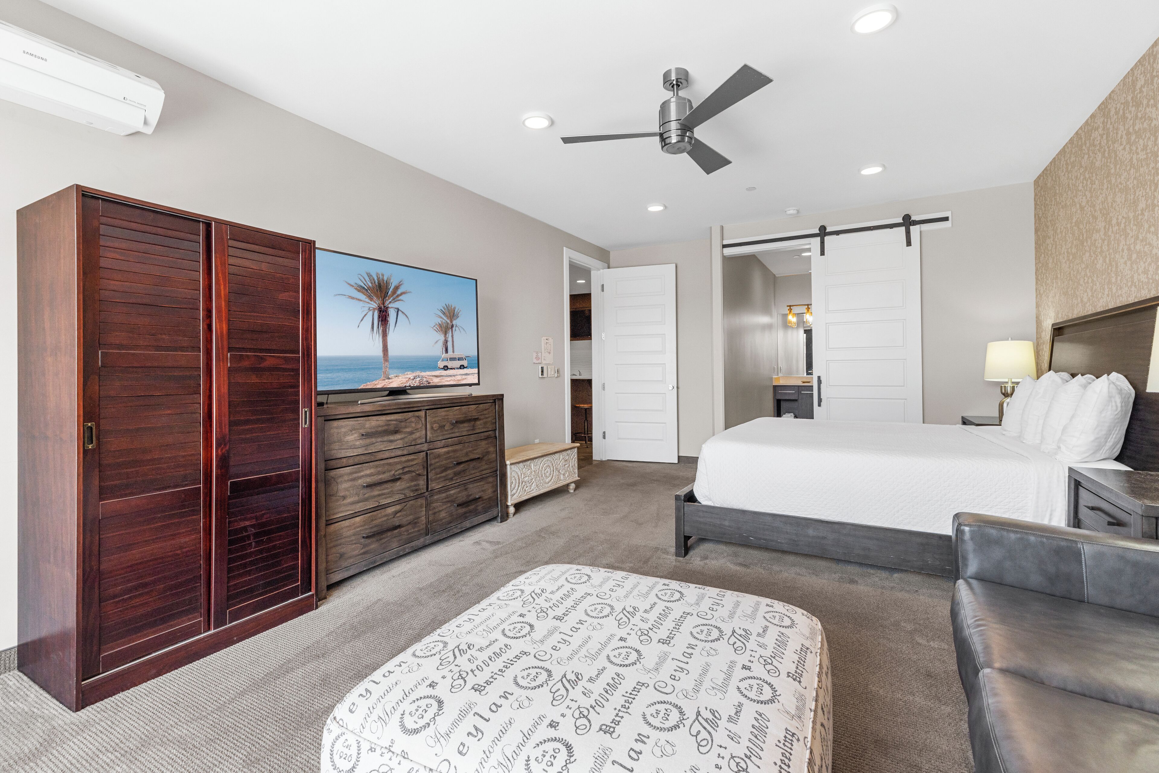 A hotel room with a bed, a desk, a wardrobe, a ceiling fan, and a painting of a beach scene.