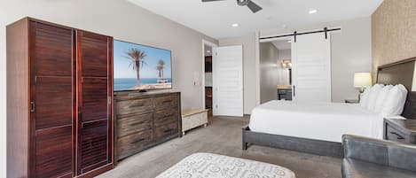 A hotel room with a bed, a desk, a wardrobe, a ceiling fan, and a painting of a beach scene.