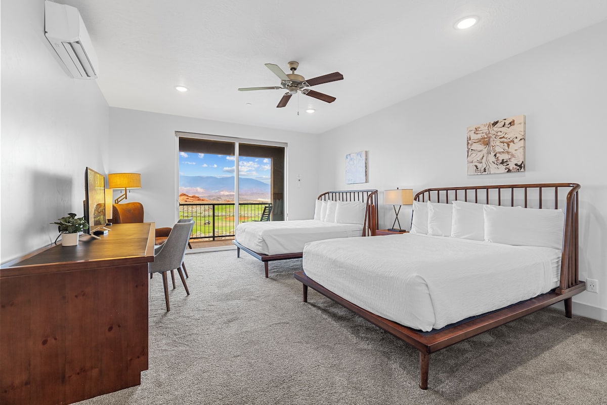 A hotel room with a desk, two beds, a ceiling fan, and a view of the outdoors.