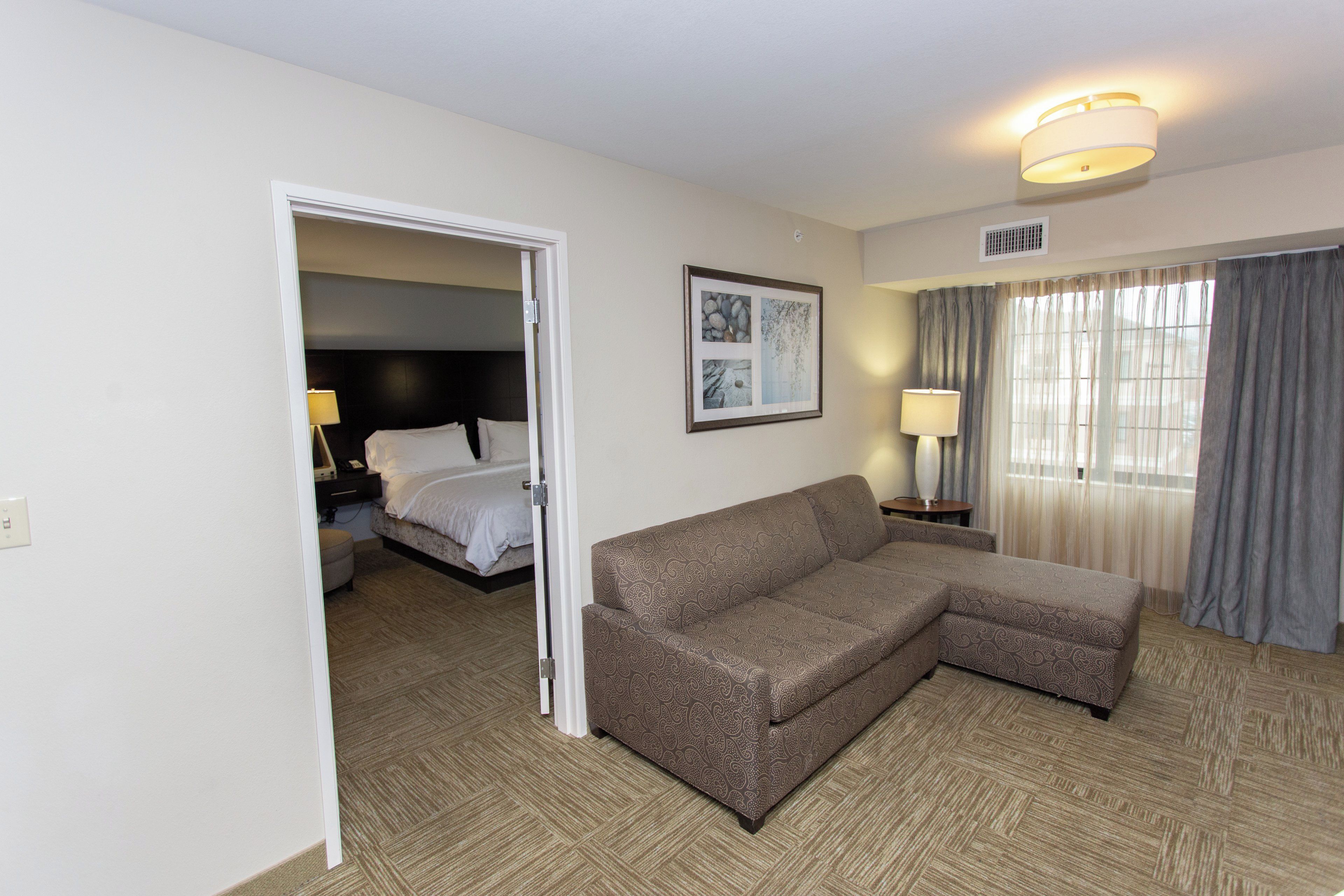 A hotel room with a sofa, a bed, a framed picture on the wall, and a window with curtains.