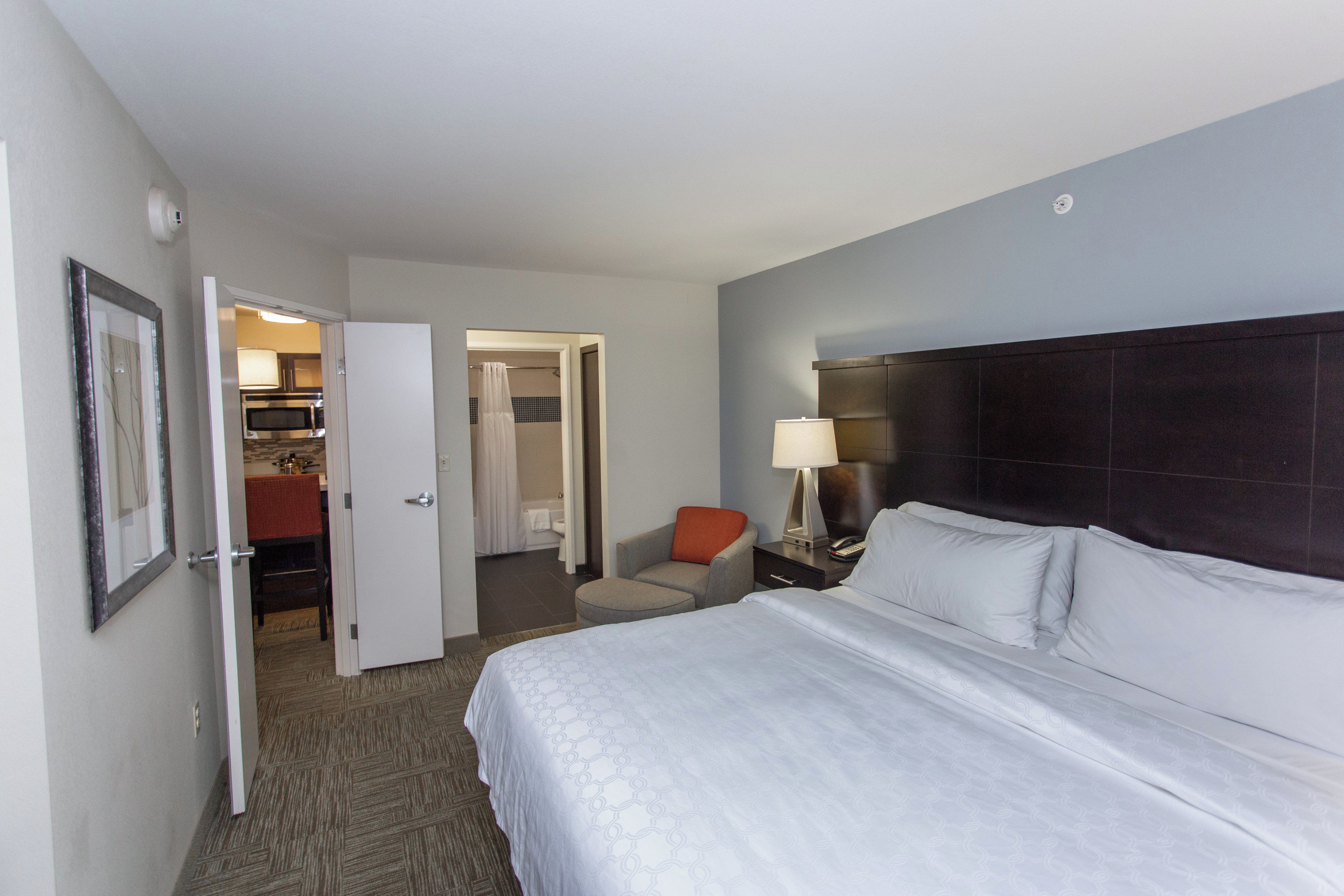 A hotel room with a bed, a nightstand, a lamp, a chair, and a bathroom visible through an open door.