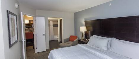 A hotel room with a bed, a nightstand, a lamp, a chair, and a bathroom visible through an open door.