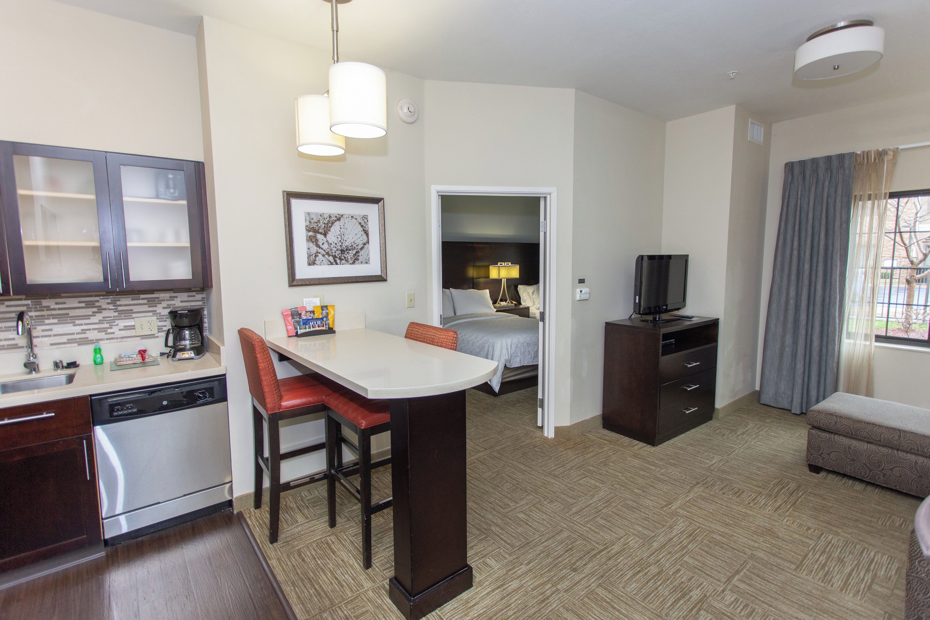 A hotel room with a kitchenette, a dining area, a bed, a TV, and a window with curtains.
