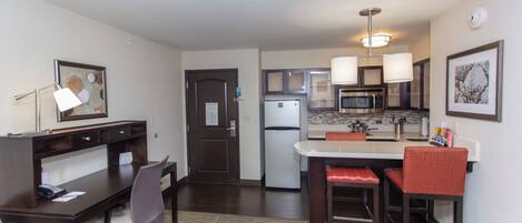 A compact hotel room with a kitchenette, a dining area with a table and chairs, a desk with a chair, and a bed.