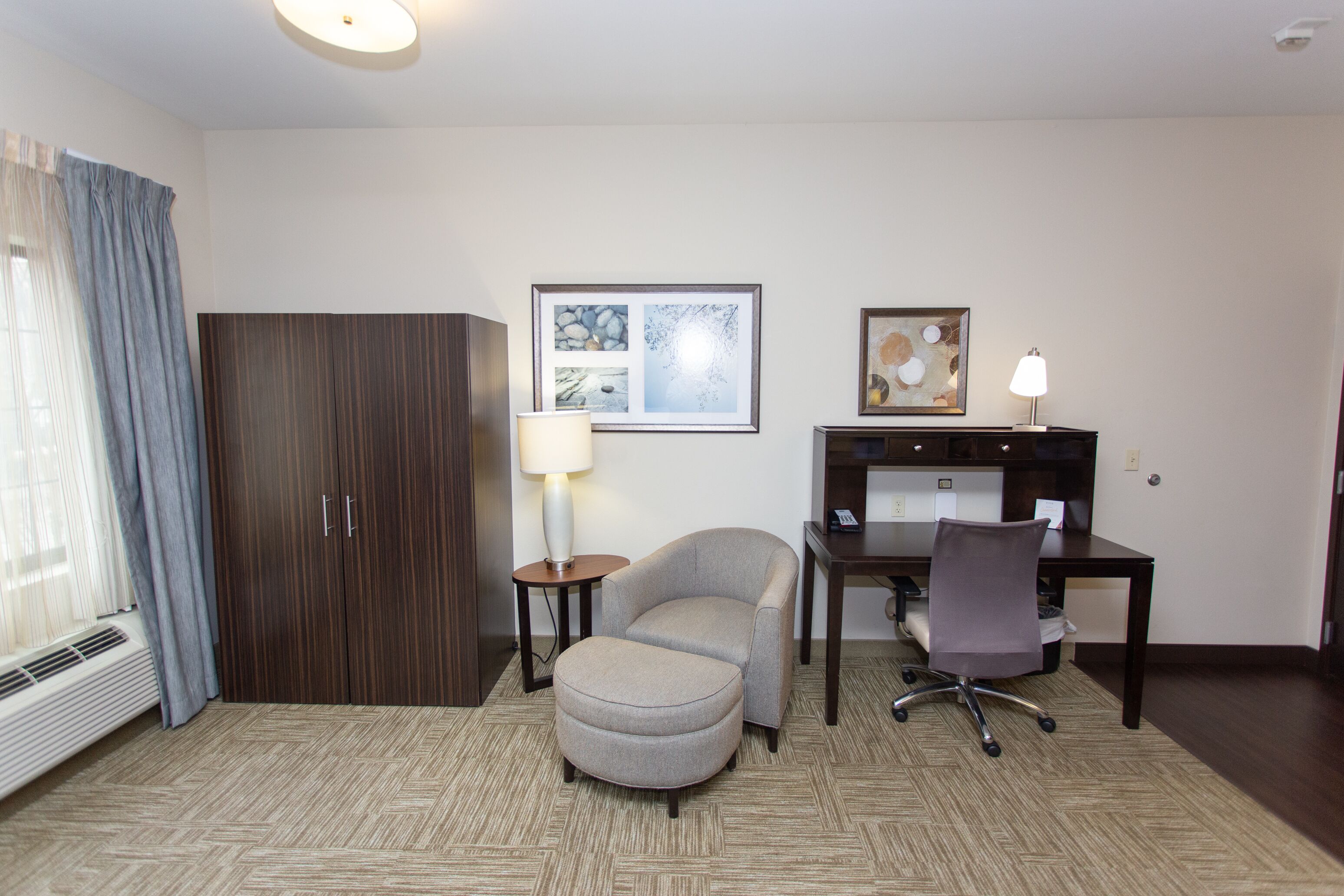 Une chambre d’hôtel avec un lit, un bureau, une chaise, une armoire, une lampe et un tableau au mur.