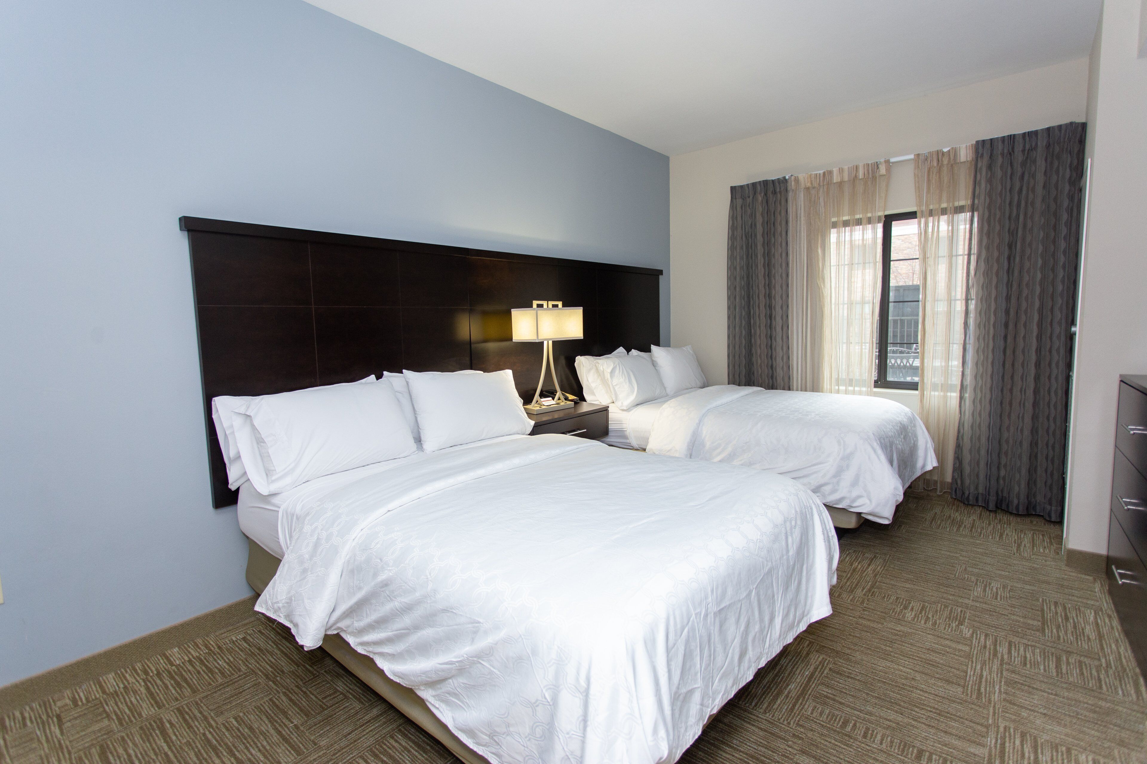 A hotel room with two beds, a nightstand with a lamp, and a view of the outside through the window.
