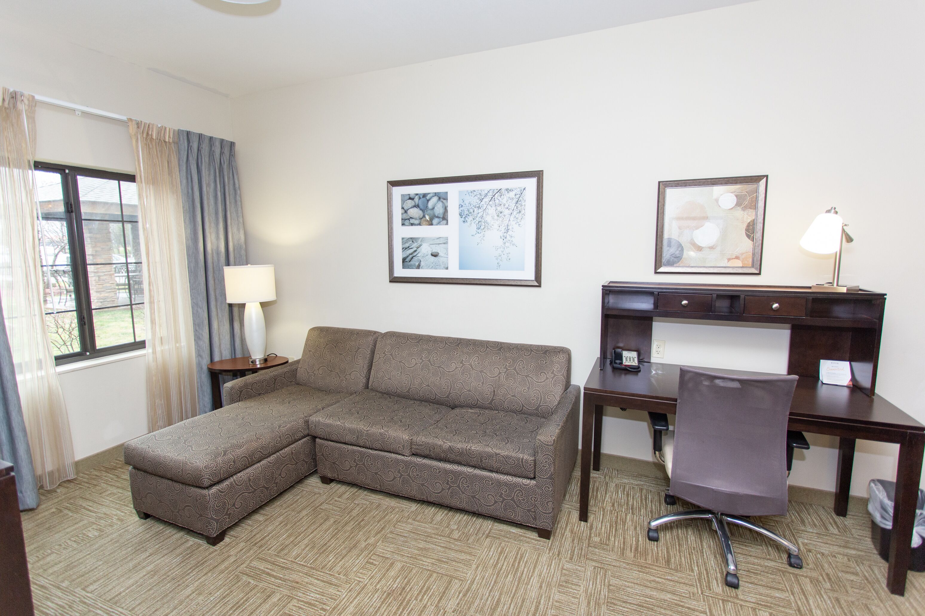 A hotel room with a sectional sofa, a desk with a chair, a lamp, and two framed pictures on the wall.