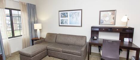 A hotel room with a sectional sofa, a desk with a chair, a lamp, and two framed pictures on the wall.