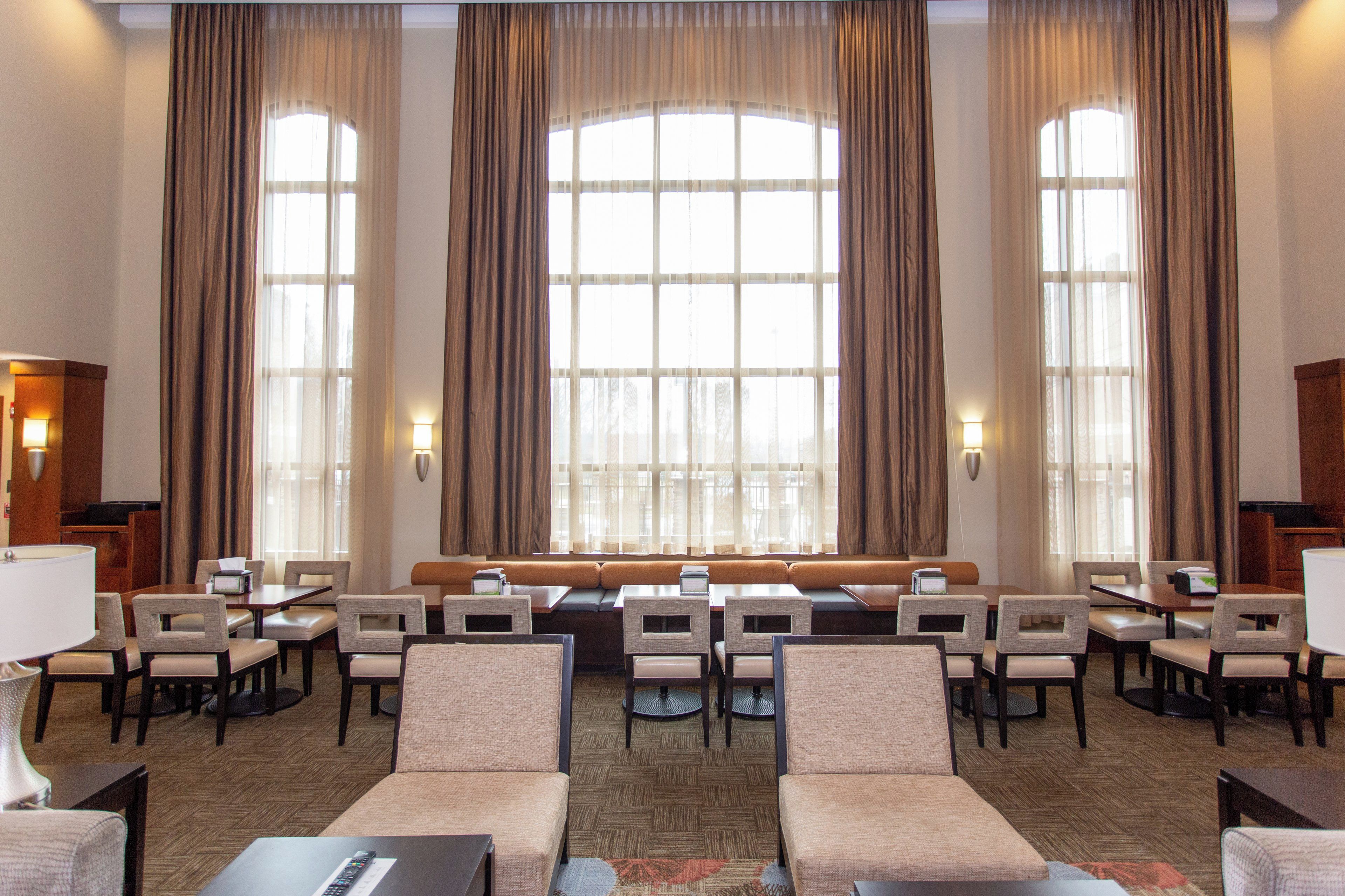 A hotel lobby with a seating area, tables, and large windows with curtains.