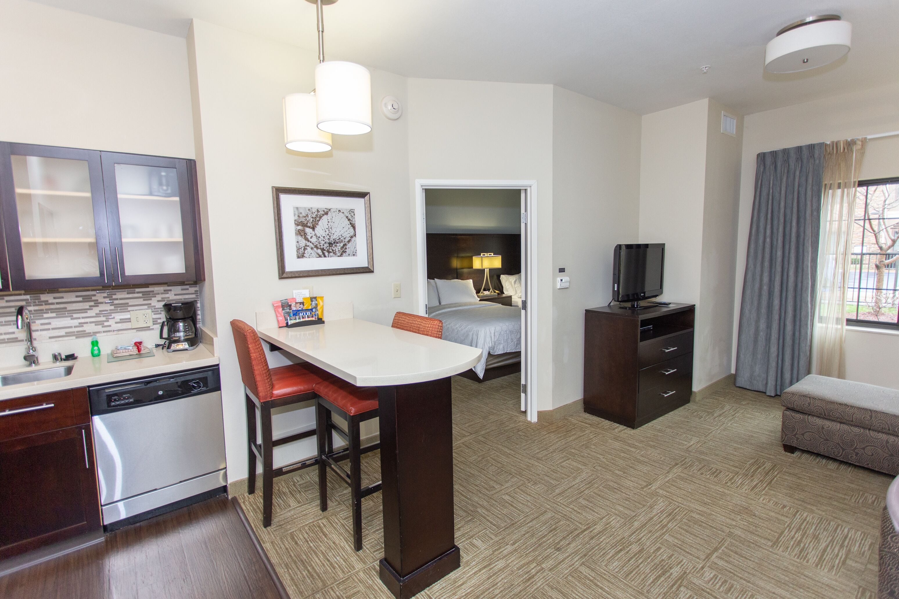 A hotel room with a kitchenette, a dining area, a bed, a TV, and a window with curtains.