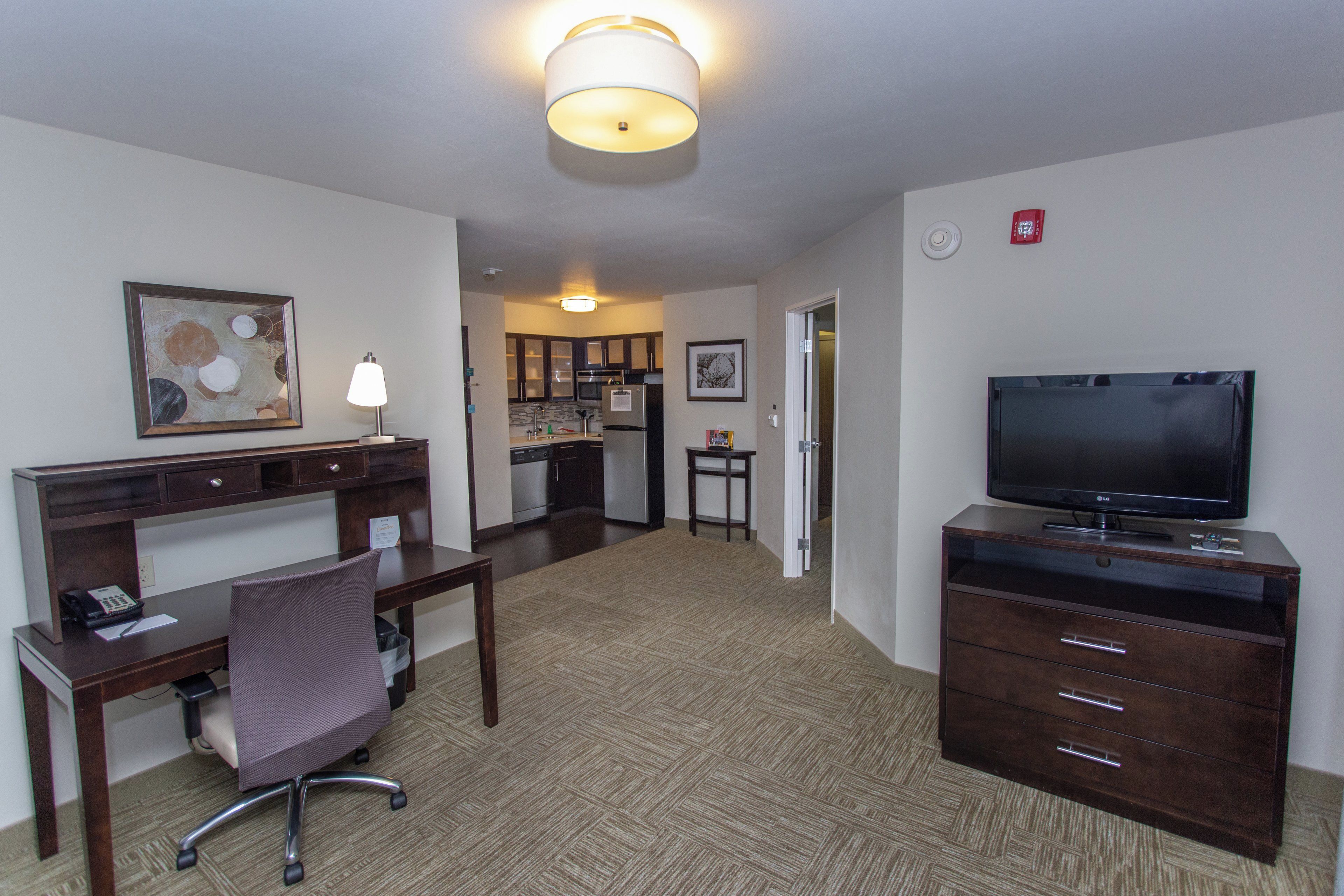 A hotel room with a desk, chair, television, kitchenette, and a hallway leading to another room.