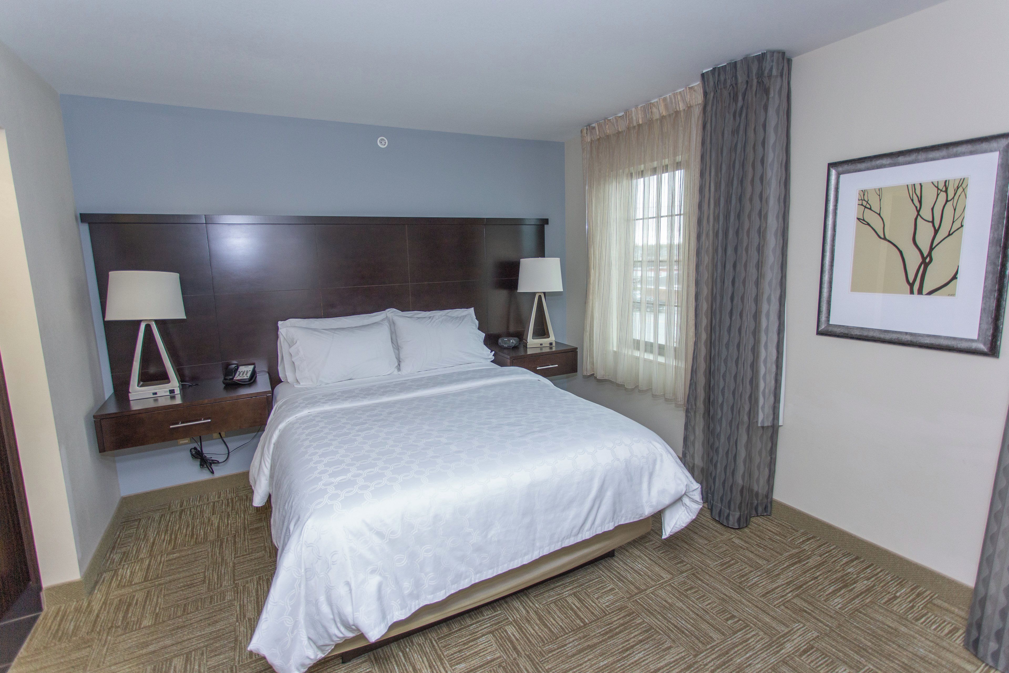 A hotel room with a large bed, two bedside tables with lamps, a window with curtains, and a framed picture on the wall.