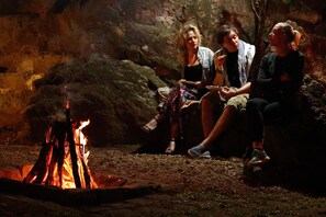 Three people sitting around a campfire in a rustic setting with stone walls.