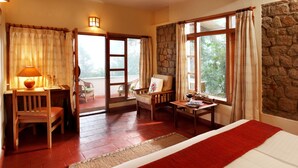 A room with a bed, a desk, and a chair. There are windows with curtains and a view of trees. - Windermere Estate (Devikolam)