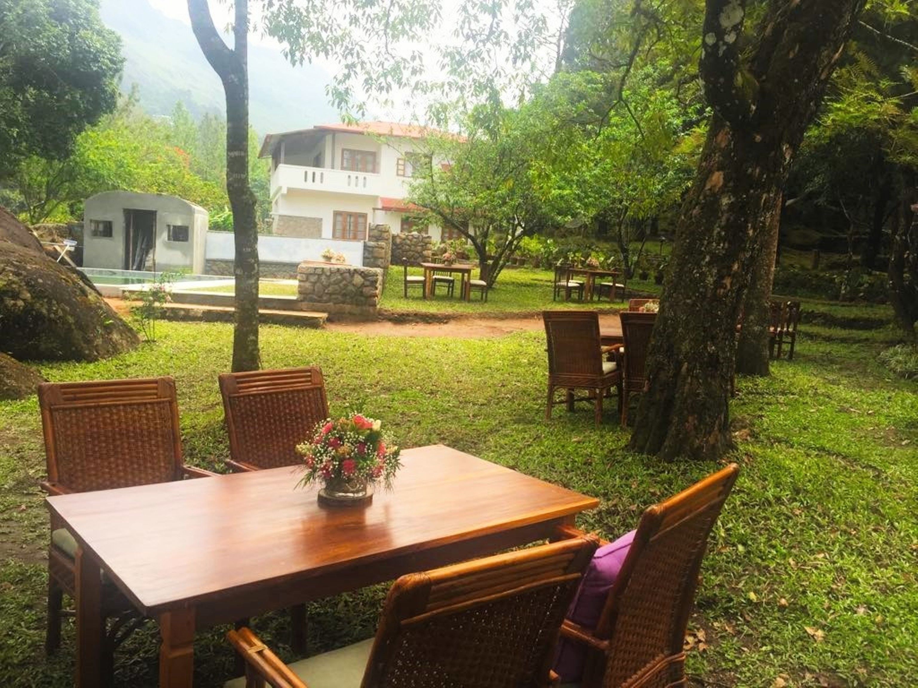 A wooden table with wicker chairs and a vase of flowers, set in a lush garden with a building in the background.