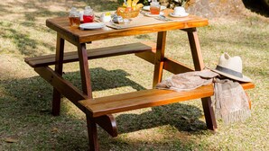 A picnic setup with a table, food, and a hat on a bench in a garden area with a building in the background. - Windermere Estate (Devikolam)