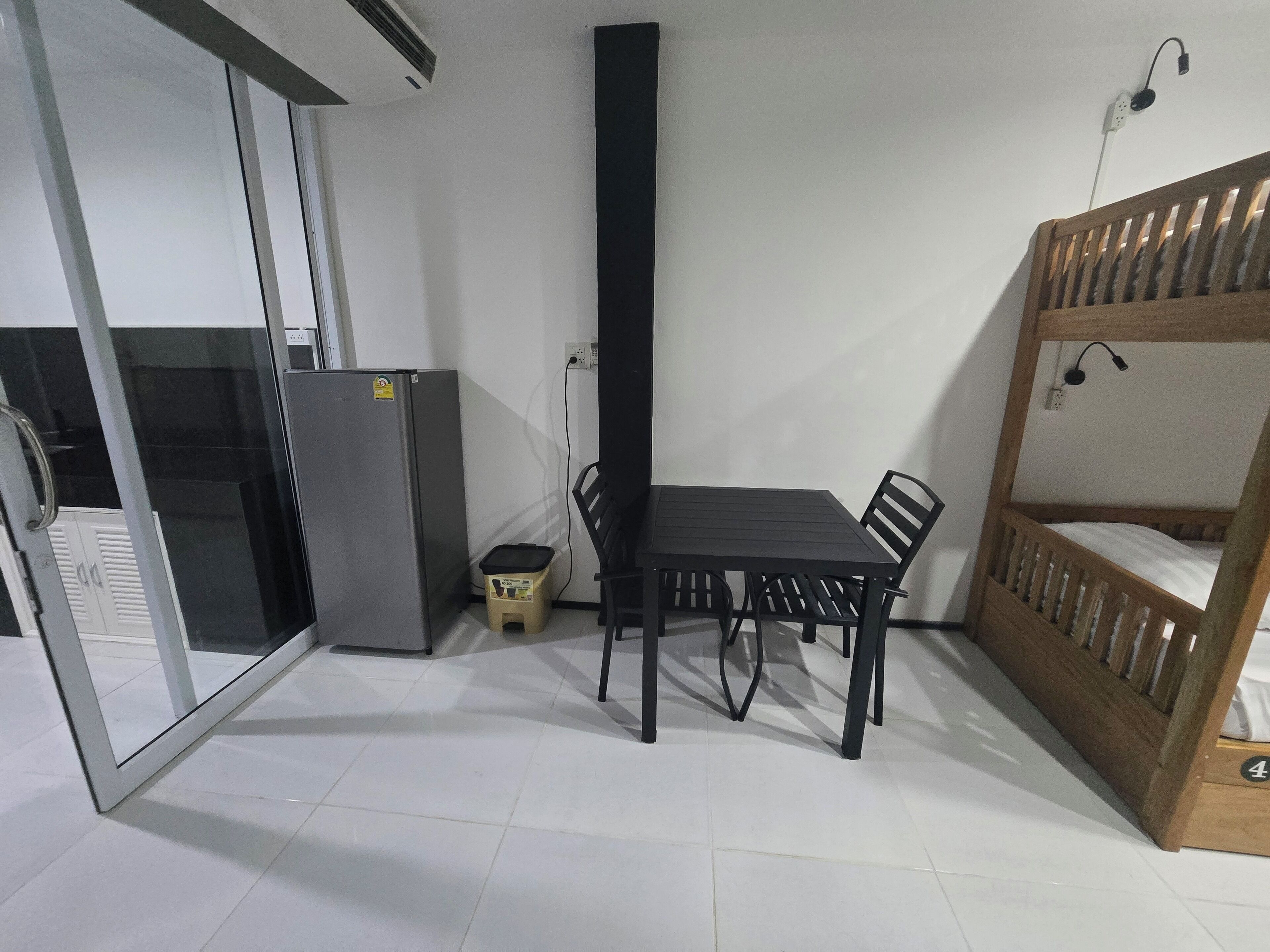 Una habitación pequeña con una mesa, sillas, una cama individual, un refrigerador y un cesto de basura.