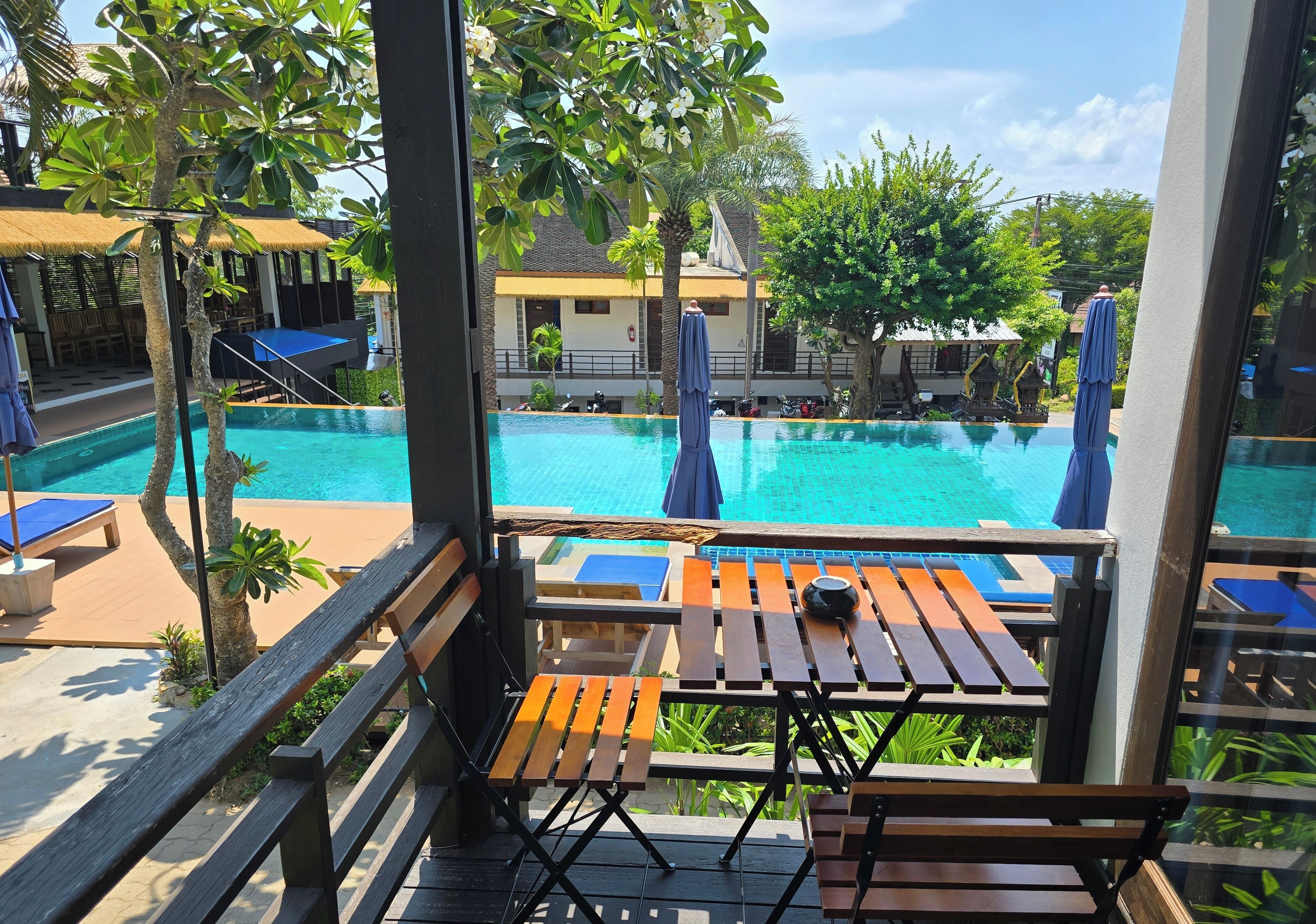 Zona de piscina con bancos y mesas de madera, una piscina azul cristalina y un edificio con techo de paja al fondo.