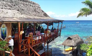 Un restaurante frente al mar con terraza al aire libre, techo de paja y vistas al océano.