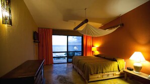 A bedroom with a bed, a nightstand, a lamp, a window with curtains, and a view of the ocean.