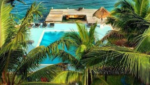 A pool area with a wooden deck, surrounded by palm trees, overlooking a clear blue ocean.