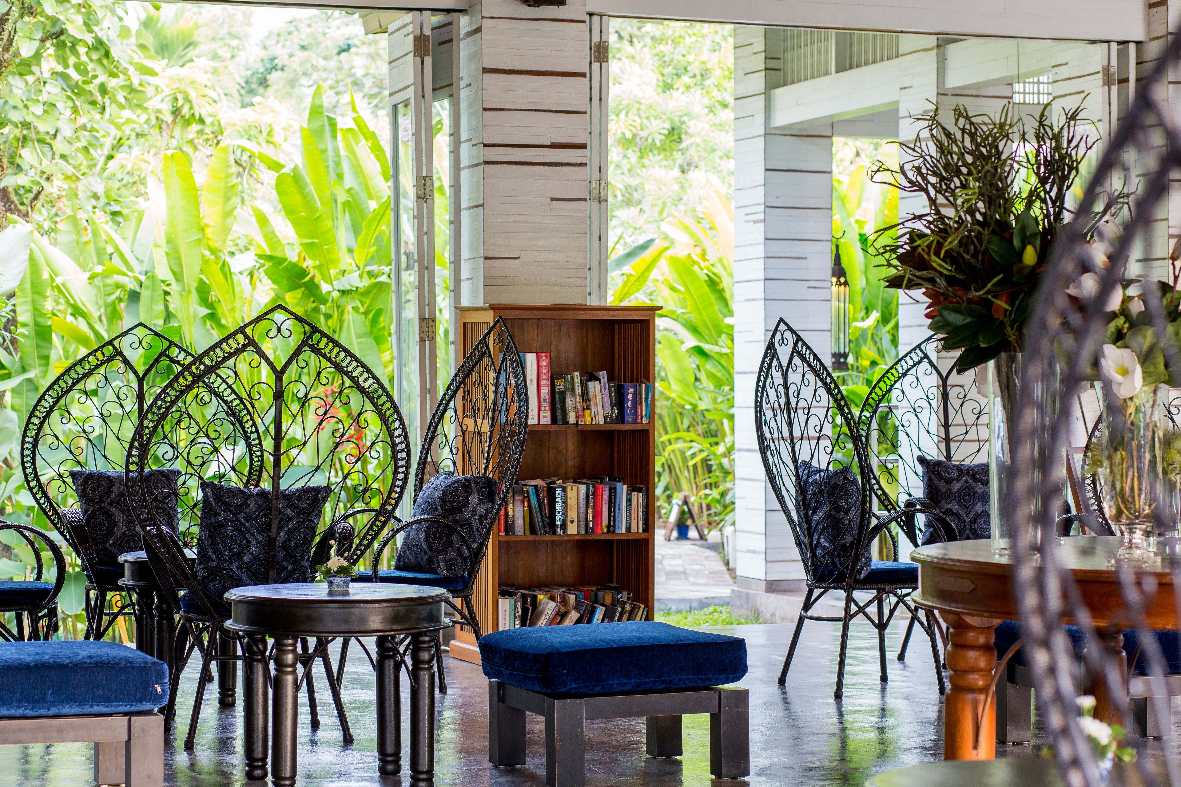 An indoor seating area with metal chairs and tables, a bookshelf, and a view of lush greenery outside.