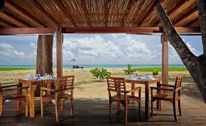 Una terraza de madera con mesas y sillas, con vista a una playa y un cielo despejado.