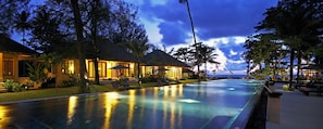 A swimming pool with lounge chairs and umbrellas, surrounded by trees and a building with a thatched roof. - Bangsak Village - Adults Only (Takua Pa)