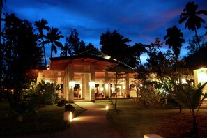 Un resort bien iluminado con un espacio exterior cubierto, palmeras y un camino que lleva a la entrada.