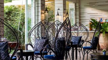 Un espace détente avec des chaises et des tables en fer forgé, de grandes fenêtres et des plantes d’intérieur.