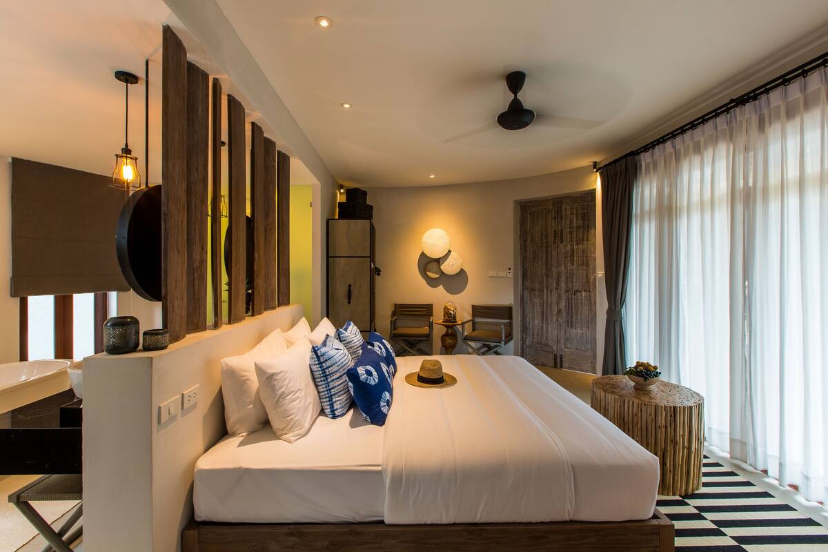 Una habitación de hotel moderna con una cama grande, cabecera de madera, televisor de pantalla plana, una mesita con un cuenco de fruta y vistas al exterior a través de cortinas de gasa.