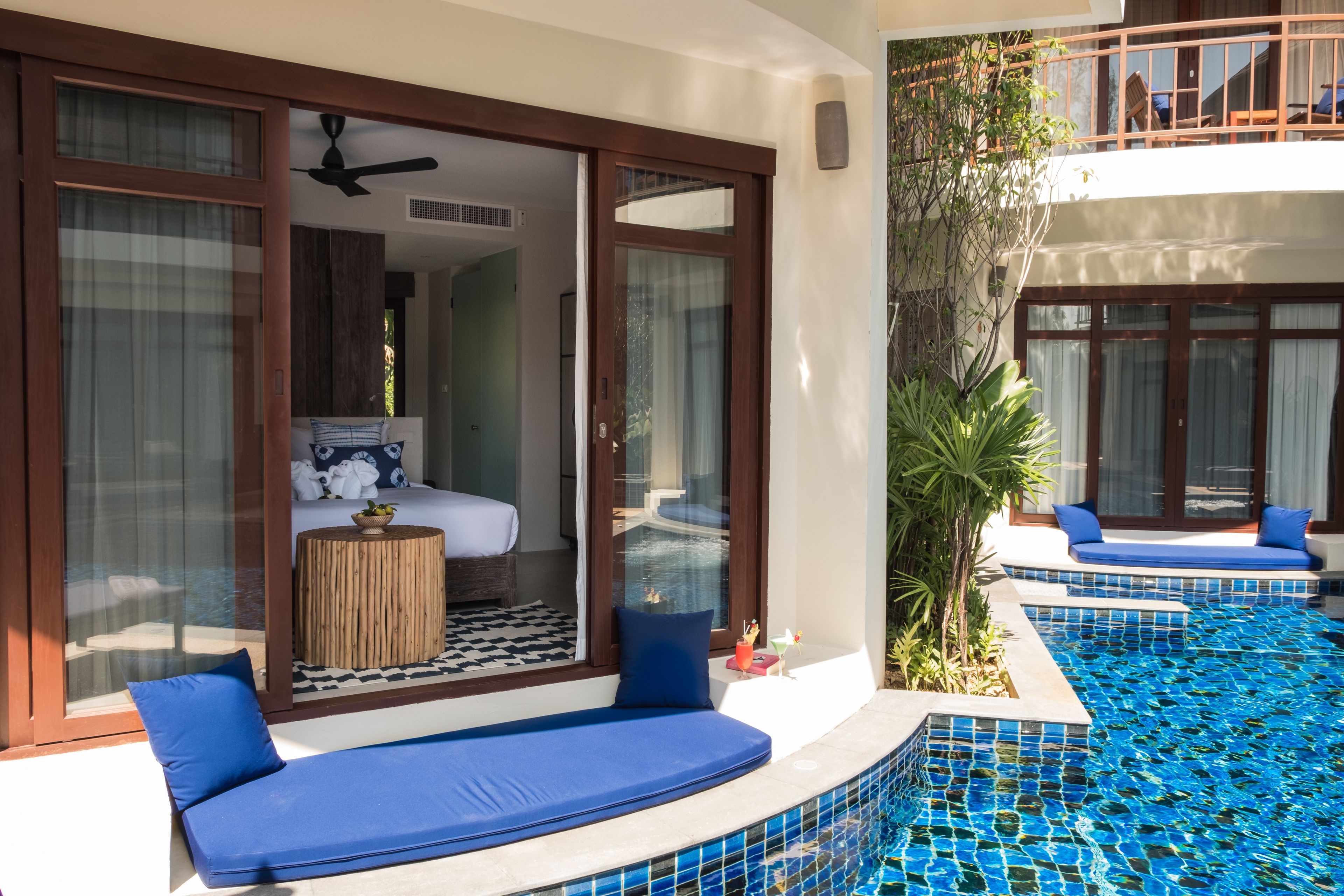 Un área junto a la piscina con una zona de estar acolchada en azul, un dormitorio con cama y una mesita, y vistas a la piscina.