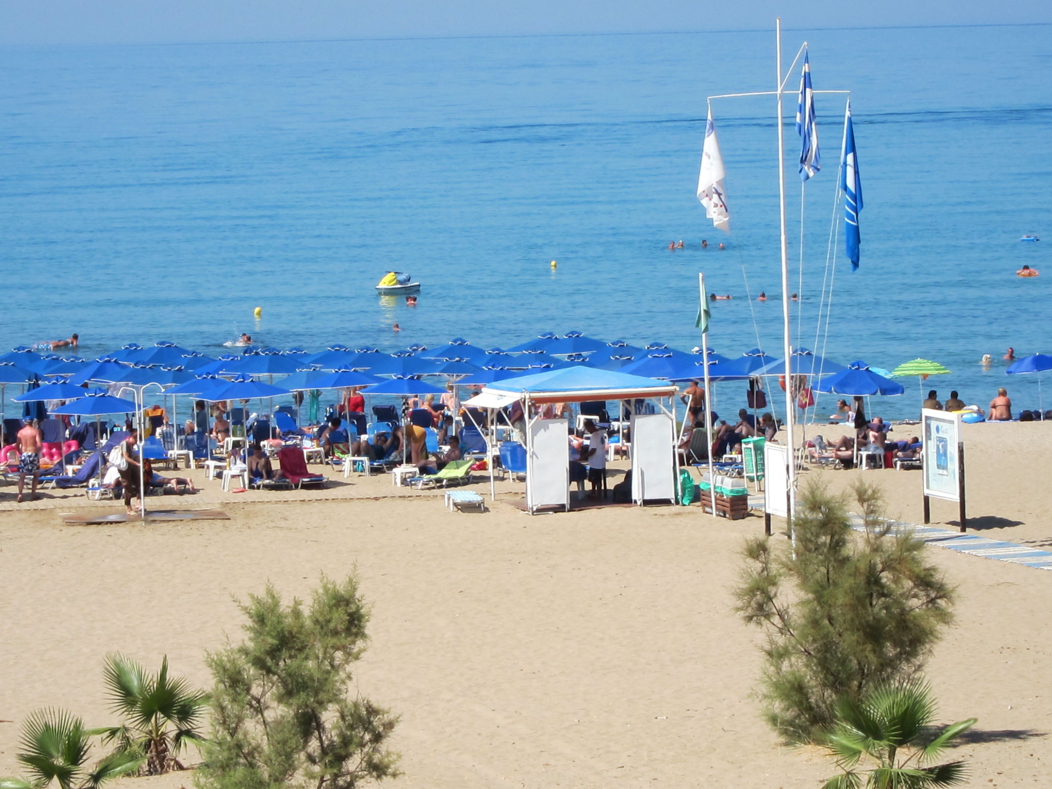 A beach with rows of blue umbrellas and beach chairs, a lifeguard station, and a clear view of the sea.