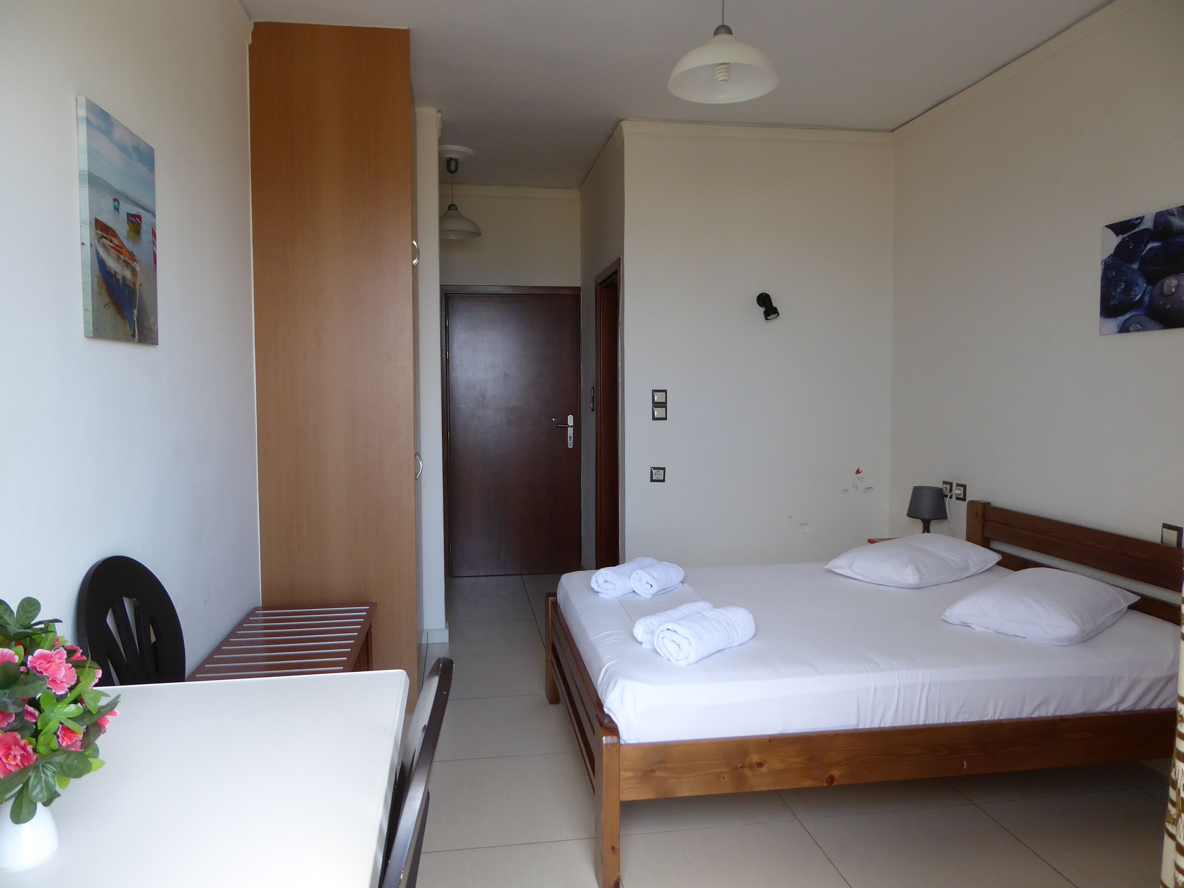 A hotel room with a wooden bed, white linens, a desk with a chair, a small table with a vase of flowers, and a painting on the wall.
