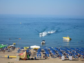 Une plage parsemée de nombreux parasols et de chaises longues, un ciel bleu azur et quelques bateaux sur l’eau.