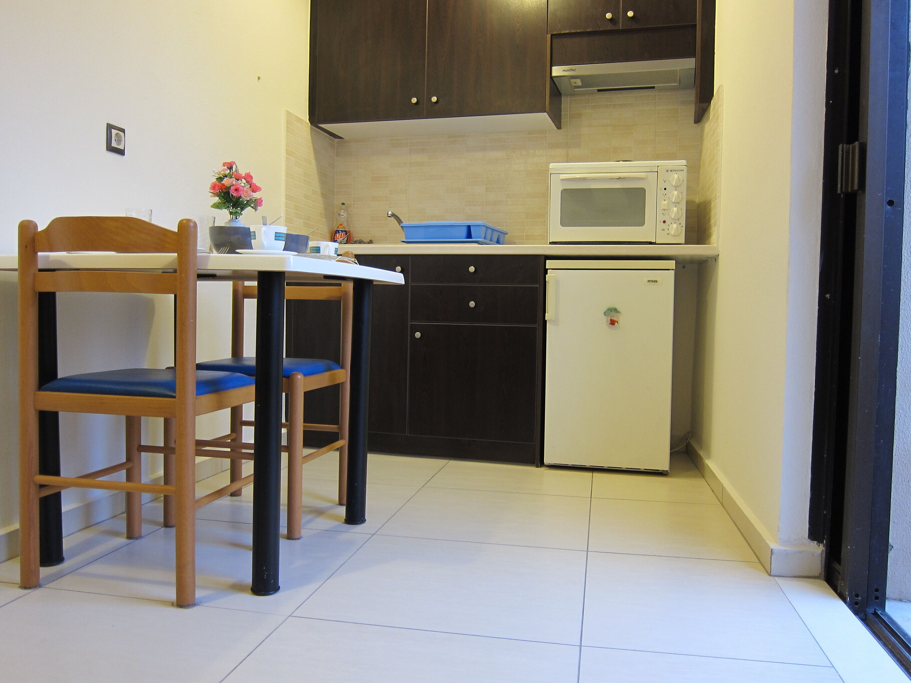 A compact kitchen with a microwave, a small refrigerator, and dark cabinetry. There is a wooden chair with a blue cushion and a small table with a vase of flowers.