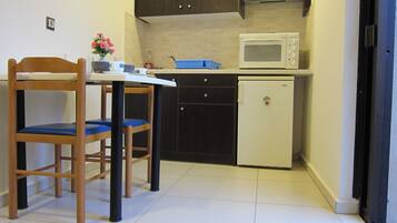 A compact kitchen with a microwave, a small refrigerator, and dark cabinetry. There is a wooden chair with a blue cushion and a small table with a vase of flowers.